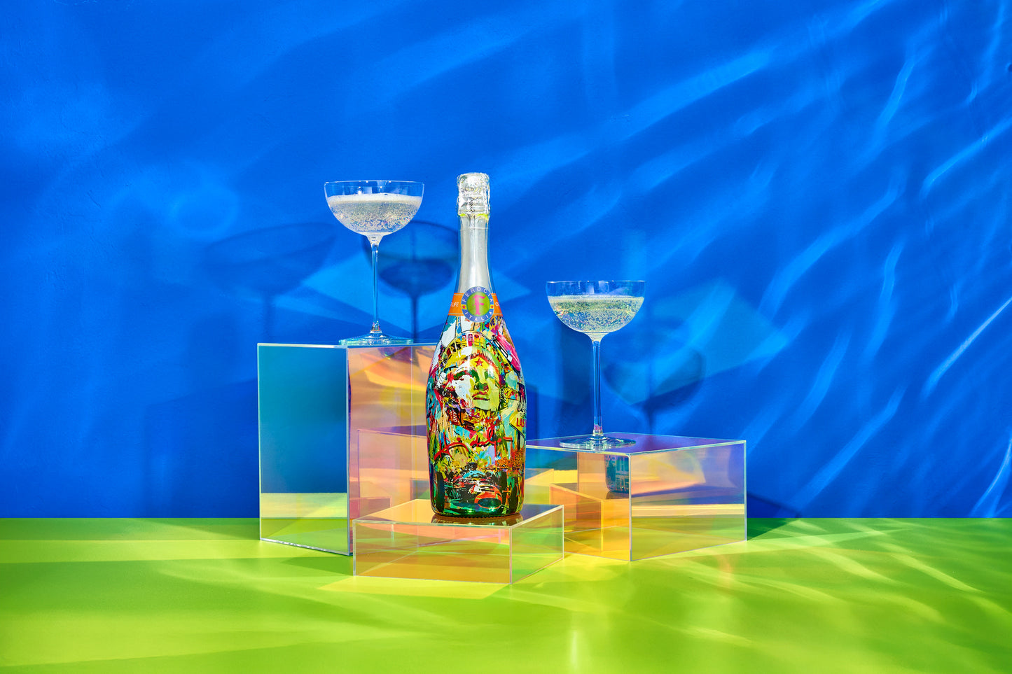 Colorful bottle with two coupe glasses on a reflective surface against a blue background.